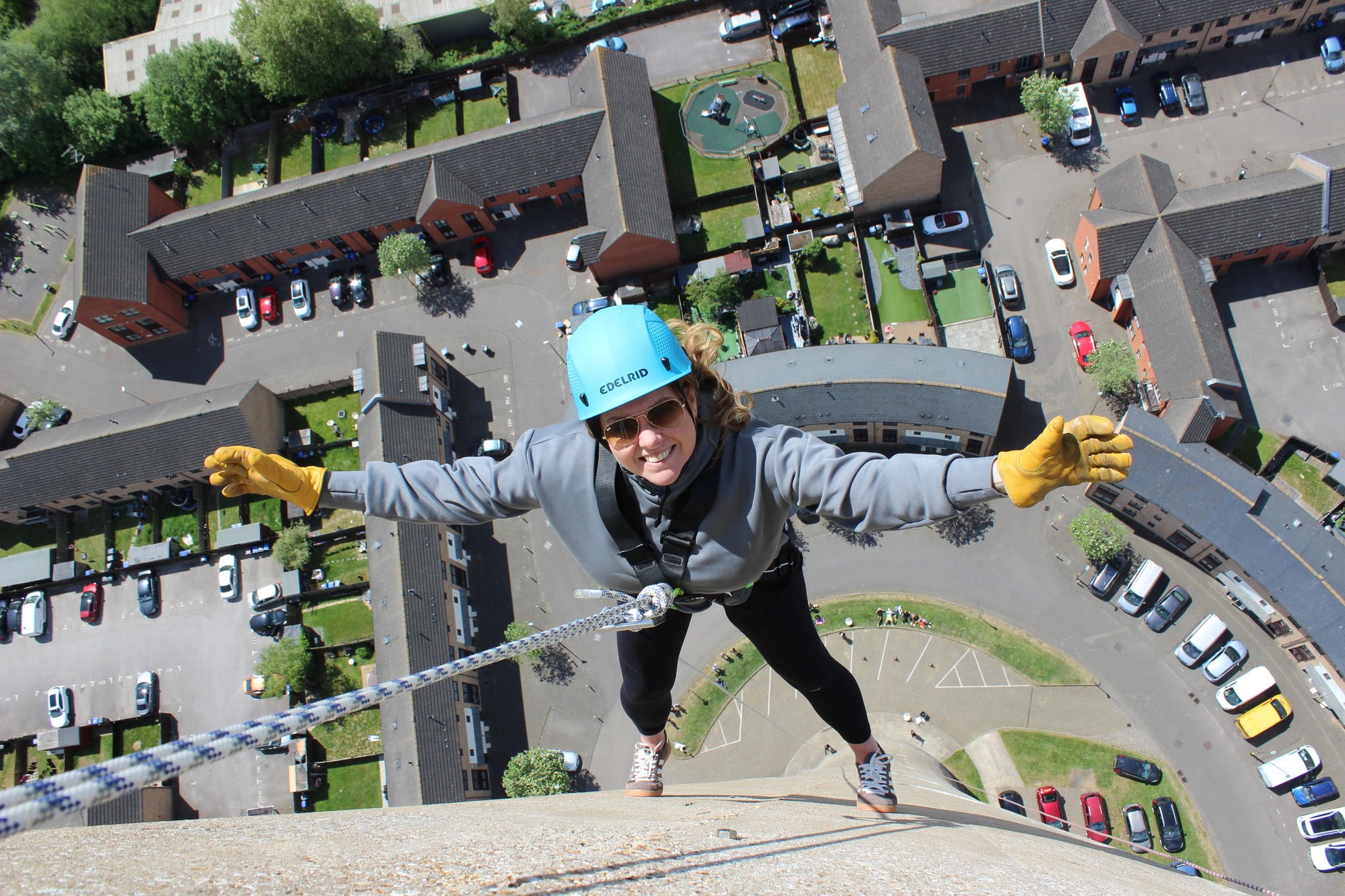 A smiling abseiler in a blue helmet with both arms outstretched high above Northampton